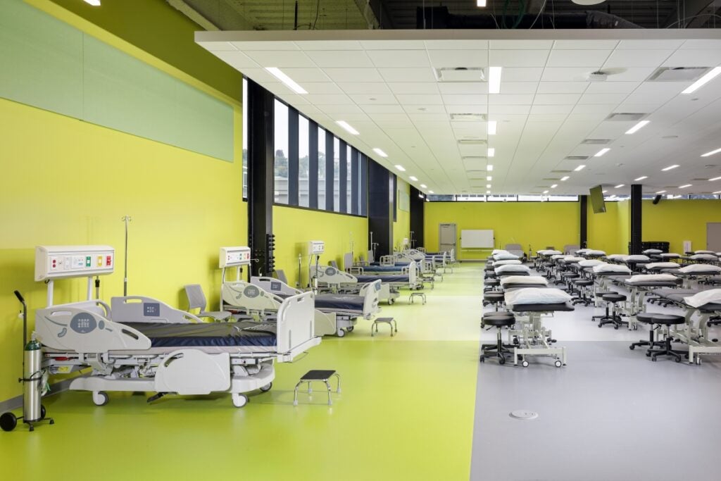 A large classroom with yellow walls has several patient tables and stools, and hospital beds lined against a wall