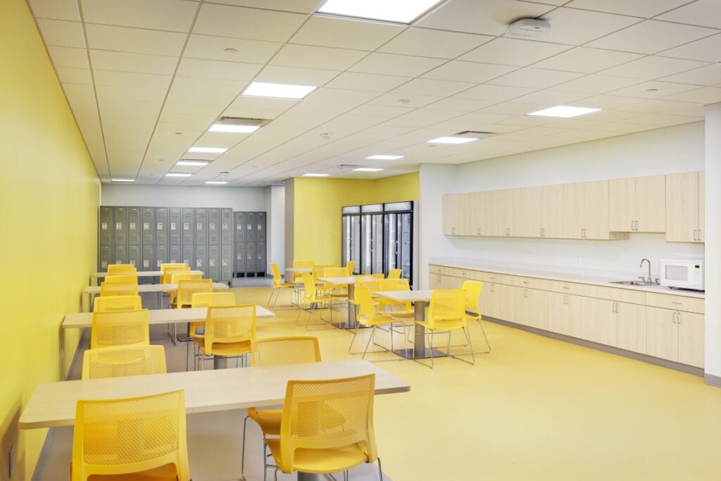 A large break room with yellow walls includes seven tables with yellow chairs, cabinets, refrigerators and lockers.