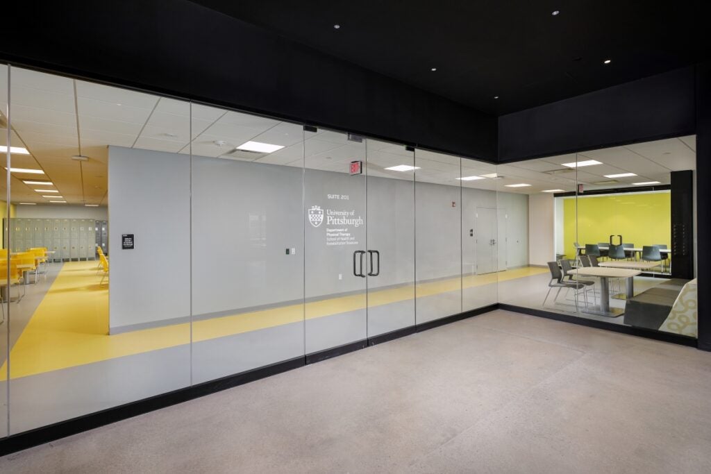 A wall of glass windows shows the interior of a building workspace including tables, chairs and lockers.