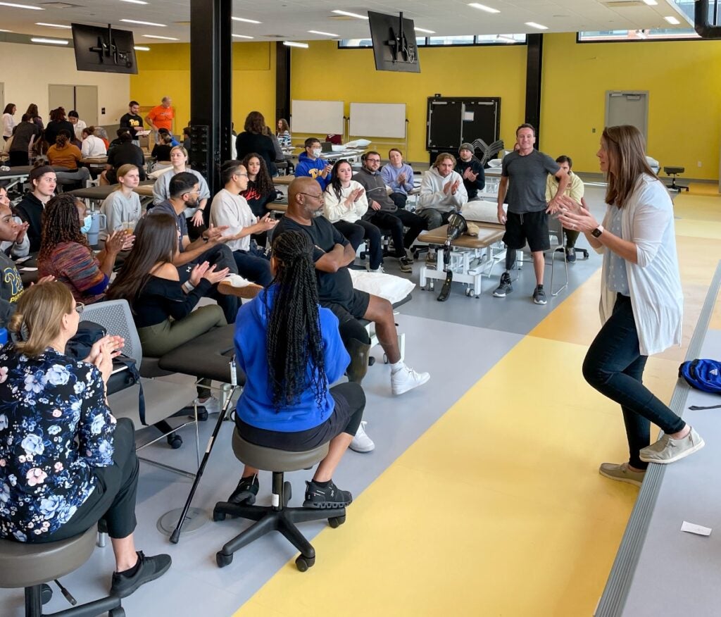 A female instructor leads a talk in front of a large group of students sitting on stools.