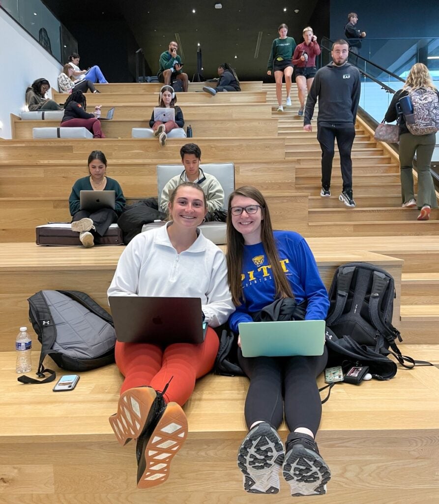 Two female students sit on a large wooden staircase with their laptops as other students study around them.