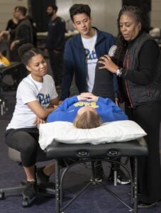 A professor demonstrates stretching a patient's leg as they lay on a table and two students watch