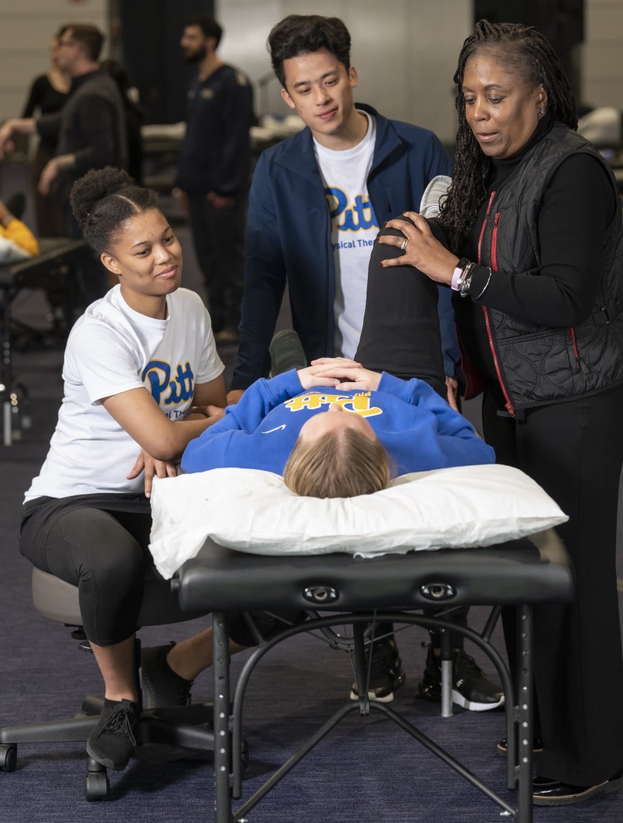 A professor demonstrates stretching a patient's leg as they lay on a table and two students watch