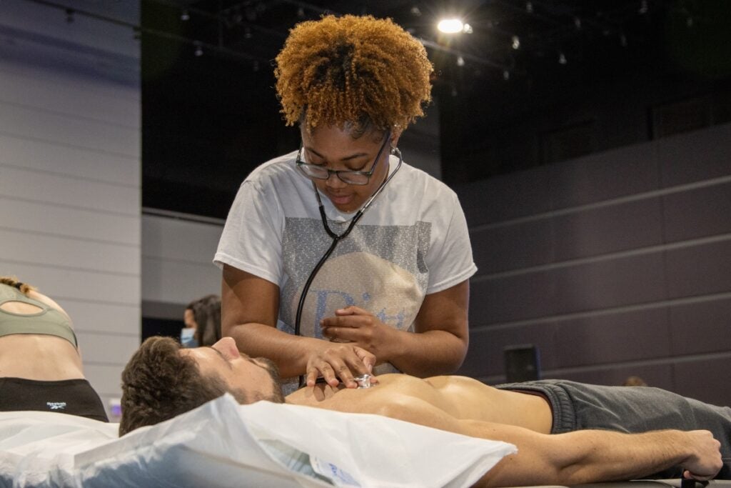 A female student uses a stethoscope to listen to the heartbeat of a male student lying on a table.