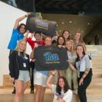 A group of 10 students pose for a photo while holding floor mats that say Pitt Physical Therapy