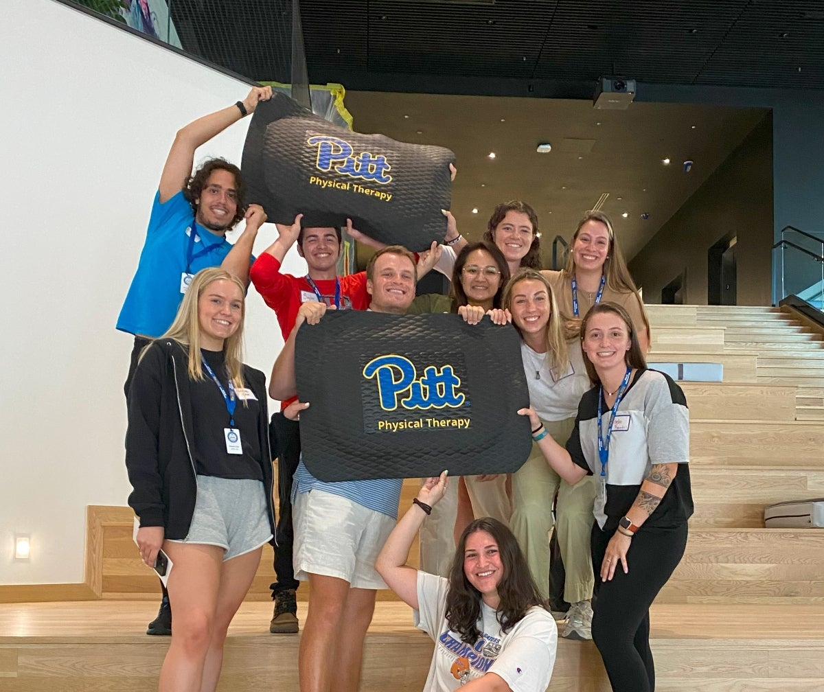 A group of 10 students pose for a photo while holding floor mats that say Pitt Physical Therapy