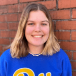 Woman with short blonde hair wearing a blue Pitt sweater and a beaded necklace in front of a brick wall.