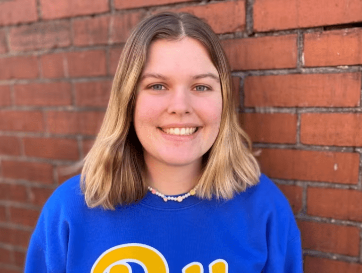 Woman with short blonde hair wearing a blue Pitt sweater and a beaded necklace in front of a brick wall.