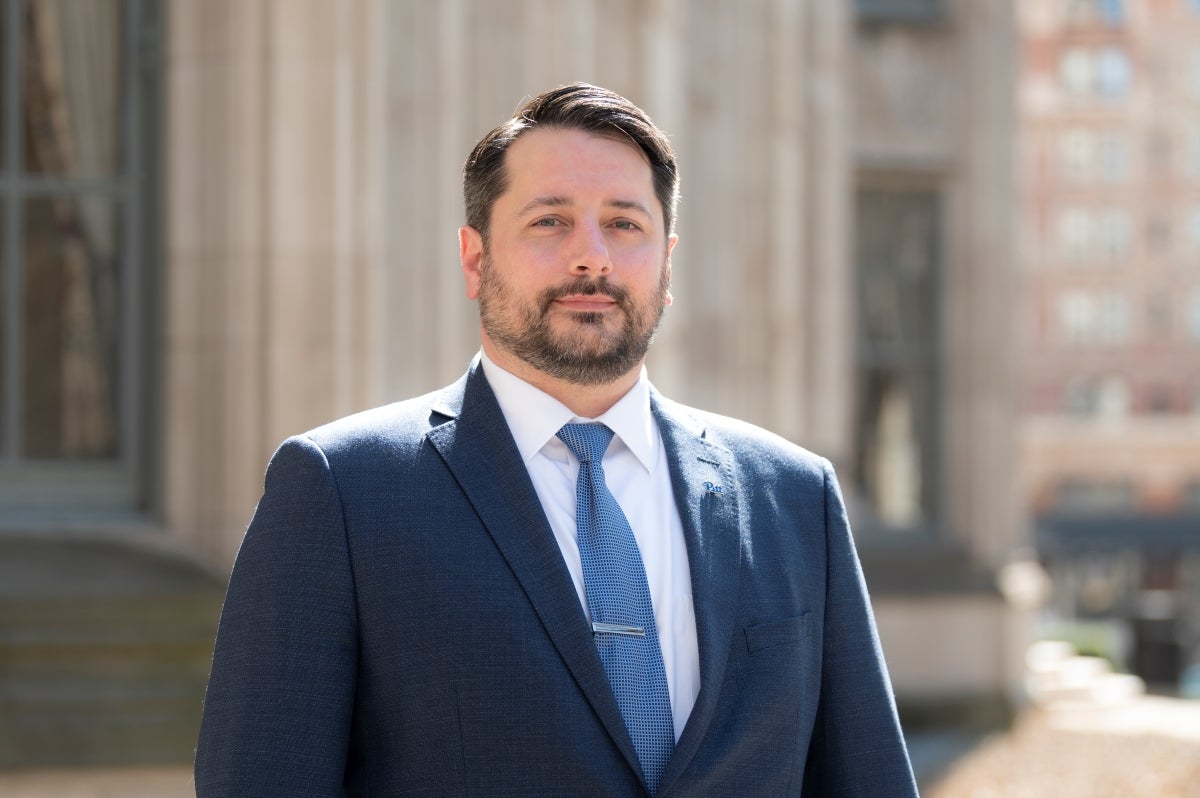 A man with short dark hair and beard wearing a dark blue suit and blue tie