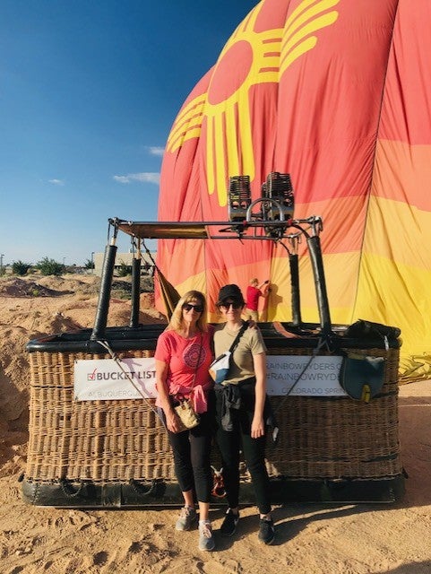 Woman with blonde hair wearing a peach shirt with a woman wearing a black hat and a green shirt standing in front of a hot air balloon