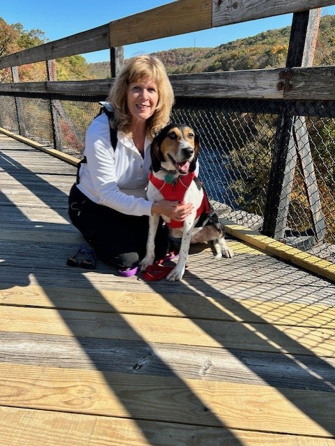 Woman with blonde hair wearing a white zip up sitting with her dog on a bridge at a state park.