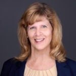 Woman with blonde auburn hair wearing a black cardigan over a tan blouse.