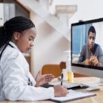 Woman with long dark braids in a white doctor coat sits at a desk taking notes while talking with a man on her computer