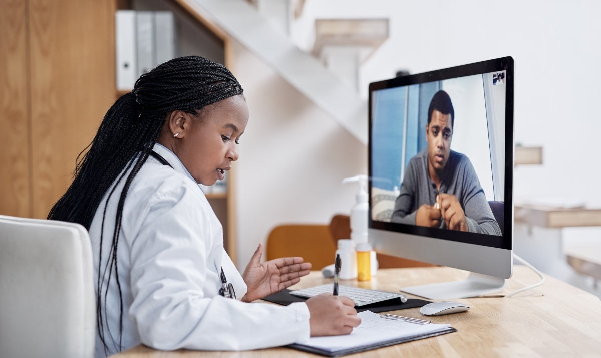 Woman with long dark braids in a white doctor coat sits at a desk taking notes while talking with a man on her computer