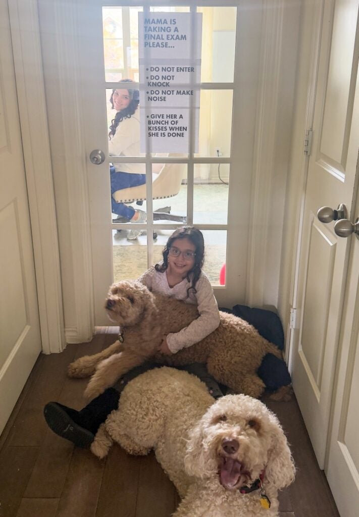 A young girl sits on the floor with two dogs while a woman smiles through the door window behind her