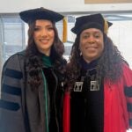 Two women in graduation regalia smile at the camera