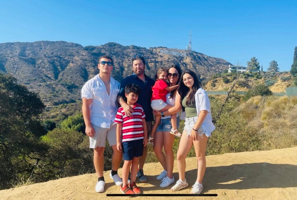 A family of six stands on a hillside with the Hollywood sign on a mountain in the background