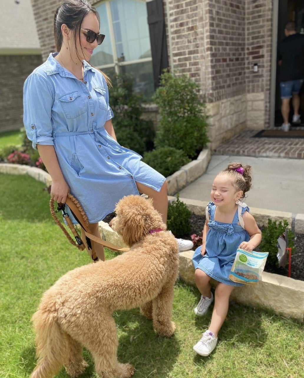 A woman in a blue dress and sunglasses smiles at a young girl sitting on a step in front of a curly haired dog