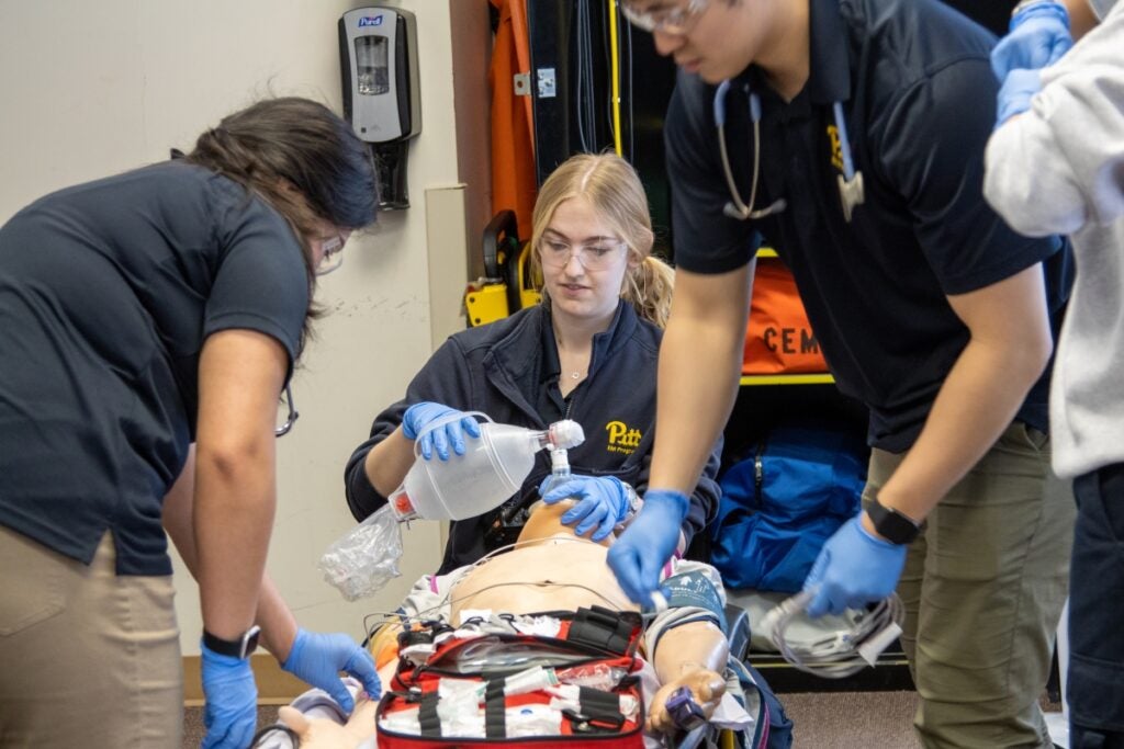 A woman with blonde hair and safety glasses holds a breathing pump to the face of a dummy patient