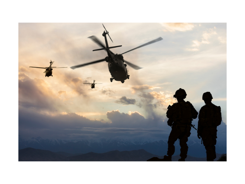 Two military personnel stand in the foreground while three helicopters fly over at sunset