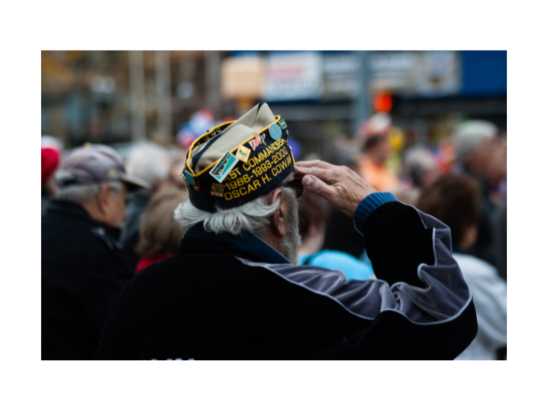 A man with gray hair and beard facing away from the camera salutes with his right arm, he wears a military hat with pins and his years of service