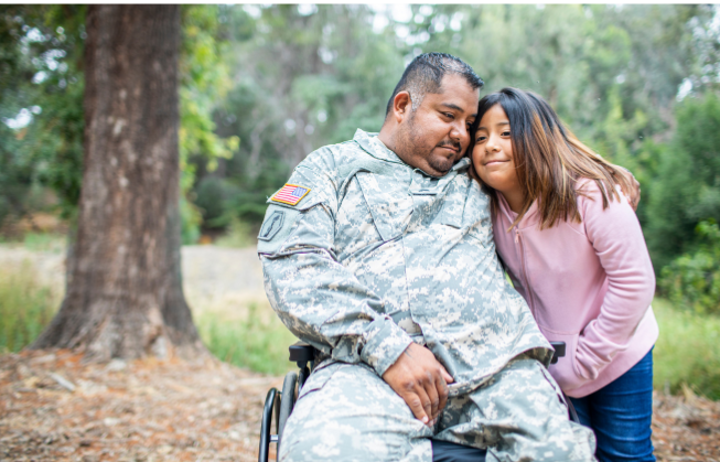 A man in military camouflage fatigues sits in a wheelchair and hugs a young girl wearing a pink sweater