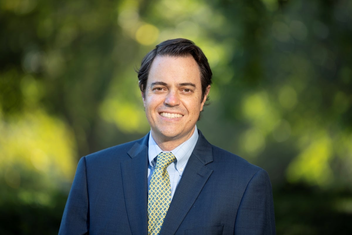 Man with short brown hair wearing a blue suit and yellow tie smiles at the camera