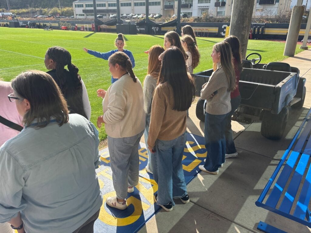 A group of young women listen to an instructor on a football field