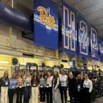 A group of women stand in a large room with exercise equipment and a large sign that says Pitt H2P