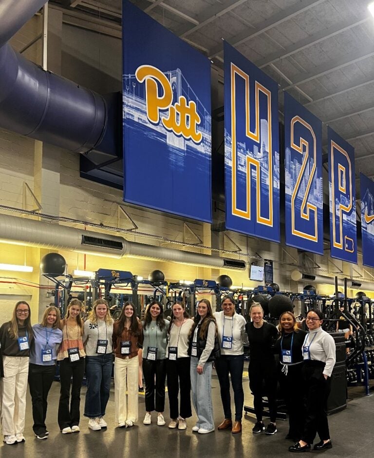 A group of women stand in a large room with exercise equipment and a large sign that says Pitt H2P