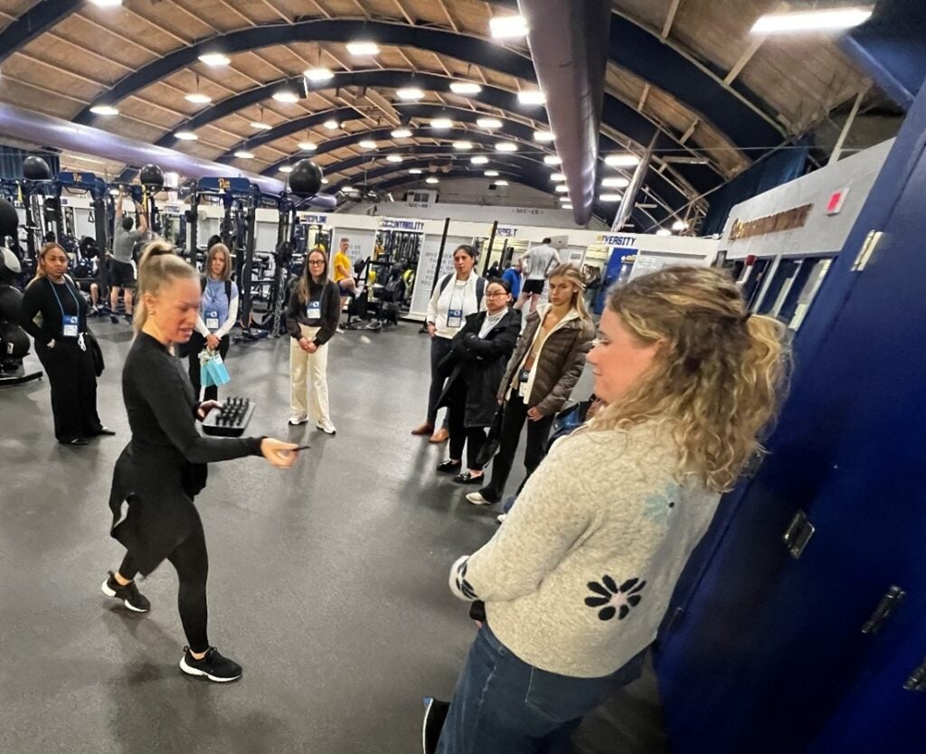 A woman with blonde hair hands out small electronic devices to a group of women in a large exercise room