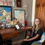 A woman with long blonde hair smiles at the camera while sitting at a desk with a laptop computer and several colorful paintings on the wall behind it