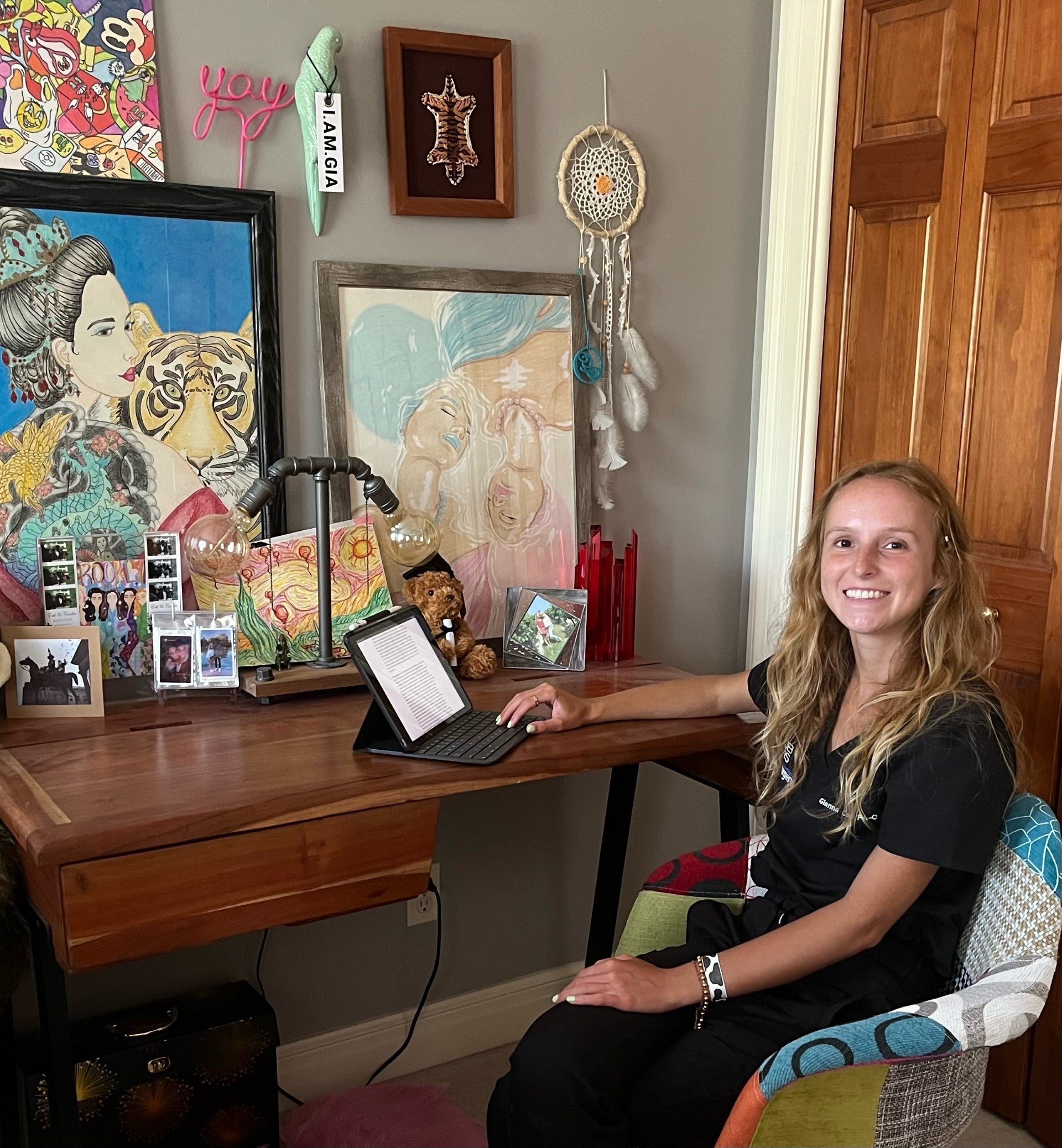 A woman with long blonde hair smiles at the camera while sitting at a desk with a laptop computer and several colorful paintings on the wall behind it