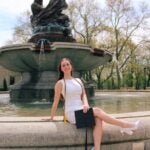 A woman in a white dress sit on the side of a fountain holding her graduation cap