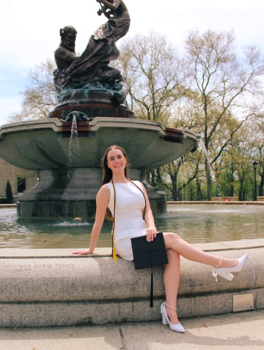 A woman in a white dress sit on the side of a fountain holding her graduation cap