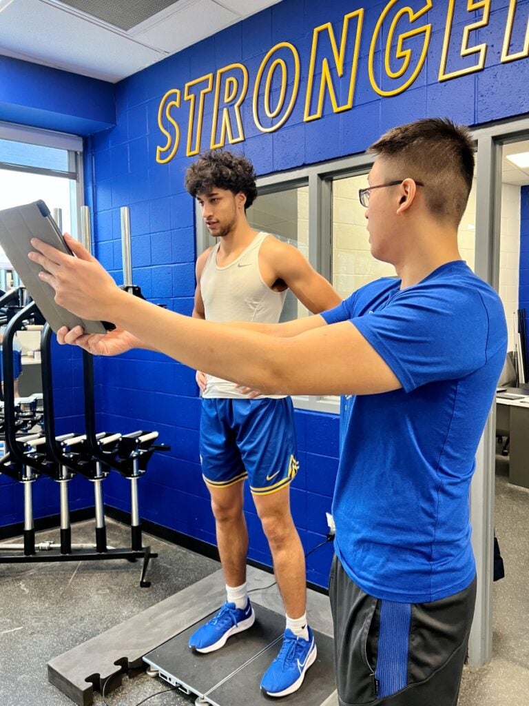 A man in a blue t-shirt and glasses holds a tablet while looking at a male athlete in a white tank shirt and blue shorts