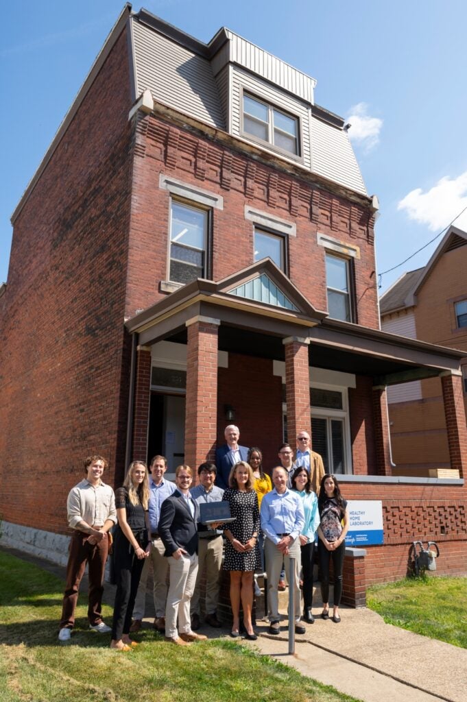 A red-bricked home with a group of 13 men and women standing in front of it.