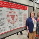 Two women smile while standing in front of a large poster of scientific information