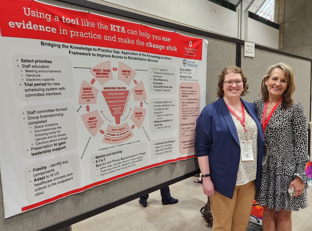 Two women smile while standing in front of a large poster of scientific information