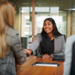 Photo of smiling woman with long dark hair reaching across a table to shake hands with a woman with long blond hair