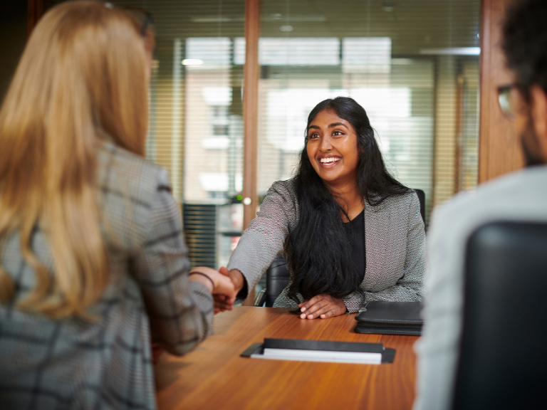 Photo of smiling woman with long dark hair reaching across a table to shake hands with a woman with long blond hair