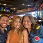 Three people in a colorful restaurant smile at the camera: a man in a polka dot shirt, a woman with long blond hair and woman with long dark hair