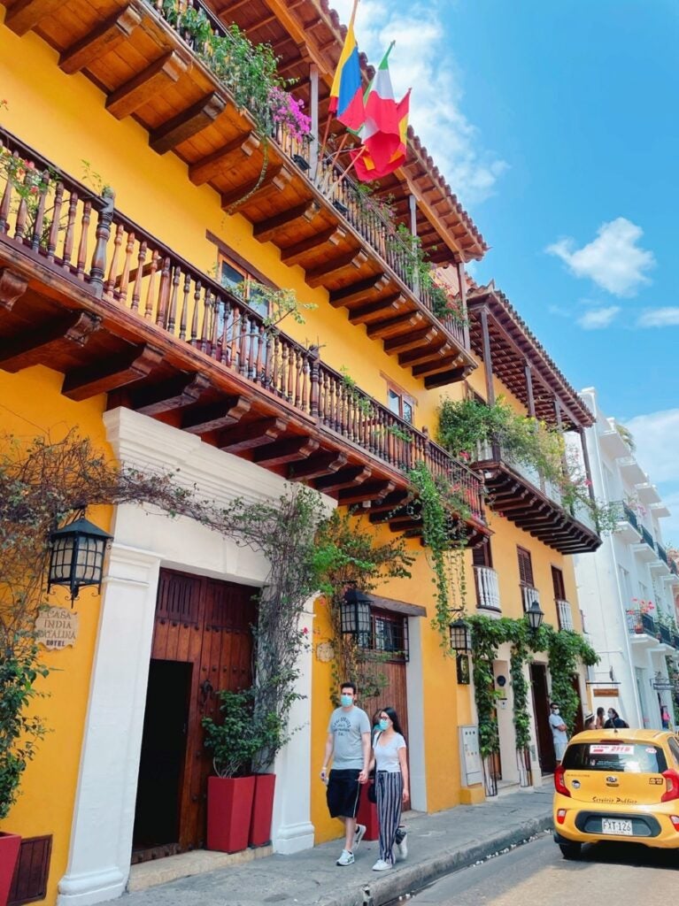 A man and woman walk on a sidewalk in front of a bright yellow building with dark wood balconies and colorful flags and flowers