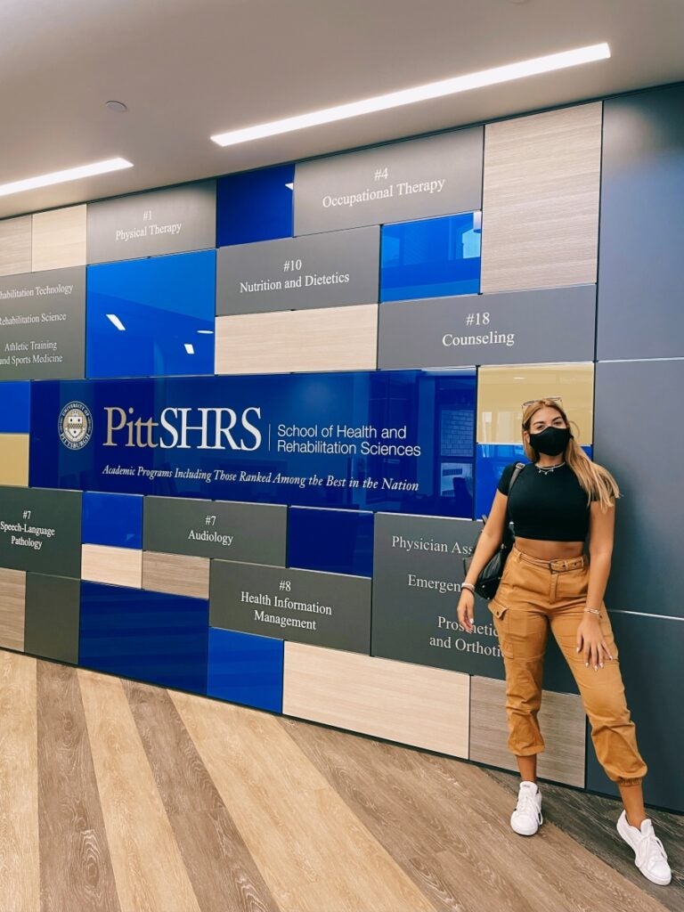 A tall woman with long blond hair wearing a black mask stands in a lobby next to a wall mosaic of large blue, yellow and gray tiles with names of different SHRS programs