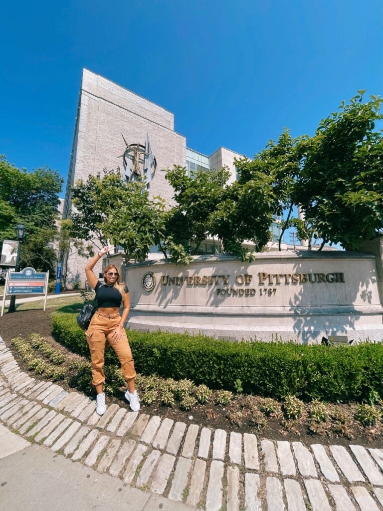 A tall woman with long blond hair waves her right hand and smiles while standing in front of a University of Pittsburgh sign