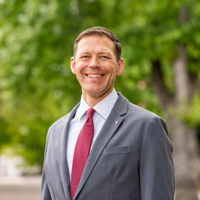 man with short brown hair in charcoal gray suit jacket and red tie smiles at the camera