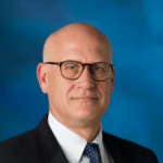 A bald man wearing a black suit jacket over a white collared shirt and a dark blue tie with dark glasses.