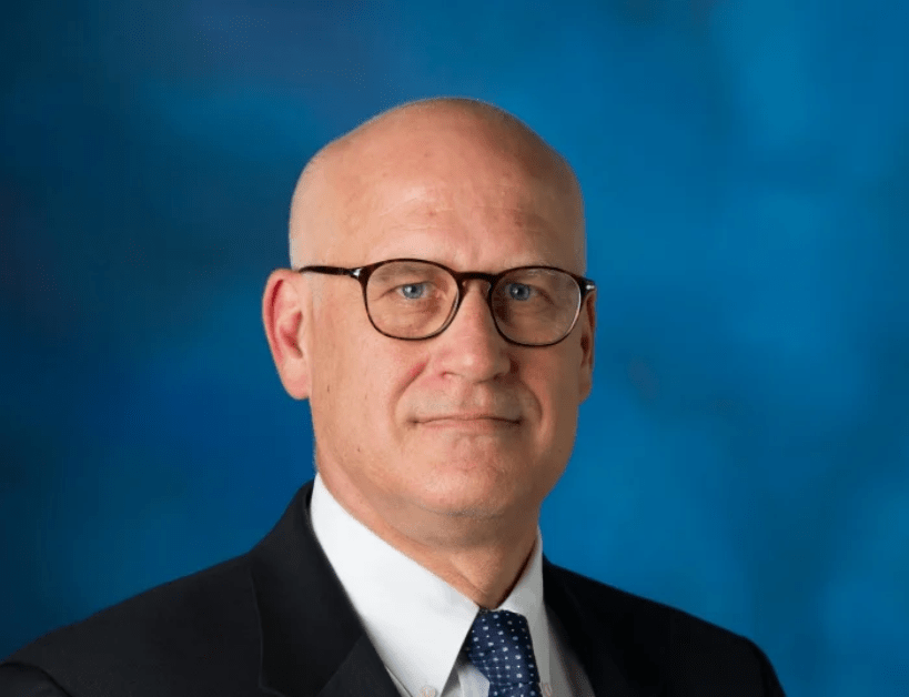 A bald man wearing a black suit jacket over a white collared shirt and a dark blue tie with dark glasses.