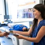 A woman with long black hair in a blue shirt smiles as she takes a clipboard from another woman