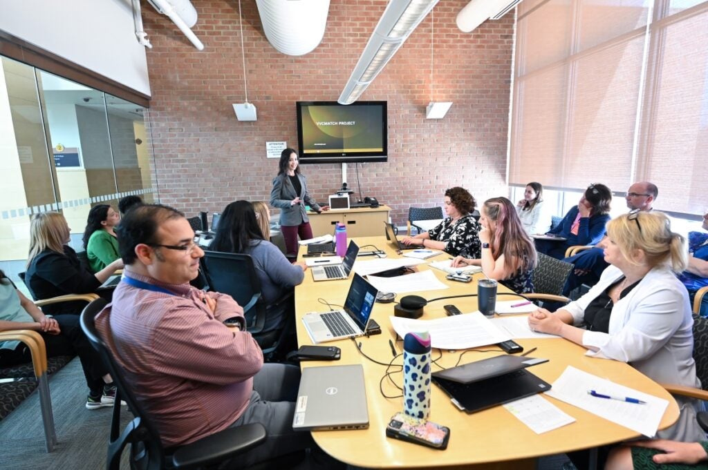 A group of people sit at a conference room table with their laptop computers while a woman stands at the front leading a discussion.